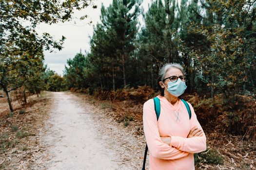 Old woman crossing arms while wearing a mask in the middle of a forest with copy space