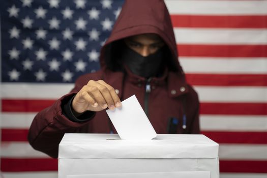 Man in hoodie with face covered casting Vote at polling booth with US falg as background - Concept of unkonwn voting or vote rigging in US elections