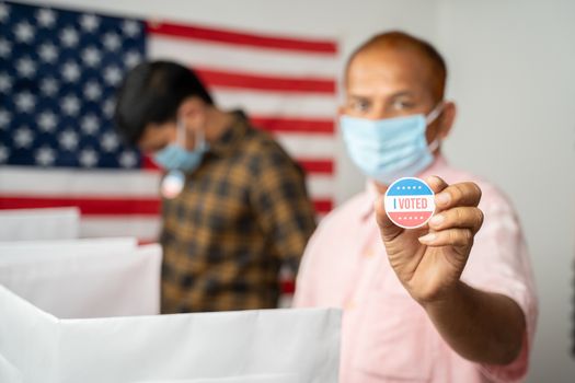 Man in medical mask showing I voted Sticker at polling booth with US flag as background - concept in person voting at US election.