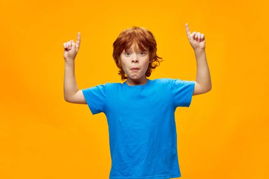 Red-haired boy grimaces face shows two hands up blue t-shirt yellow background