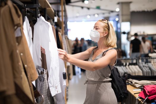 Fashionable woman wearing protective face mask shopping clothes in reopen retail shopping store. New normal lifestyle during corona virus pandemic