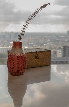 Dried flowers in beautiful ceramic red vase handmade with Woven rattan traditional box on marble table of living room and city view. Home decor. Space for text. Selective focus.