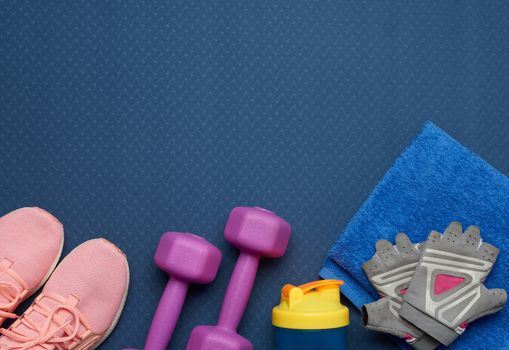 pair of purple dumbbells, sports gloves and pink gym shoes on a blue neoprene mat, top view
