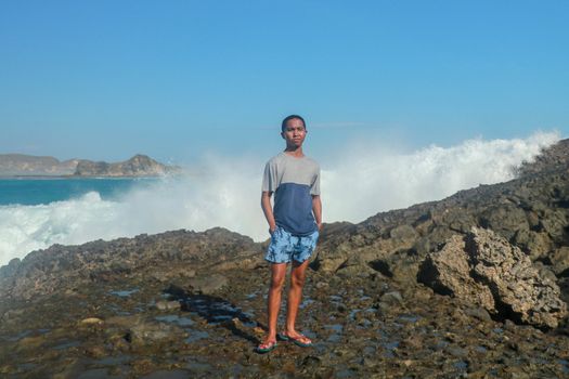 Waves hitting round rocks and splashing. A young man stands on a rocky shore and the waves crash against a cliff
