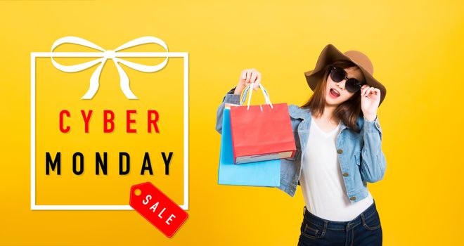 Asian happy portrait beautiful young woman smiling stand with sunglasses excited holding shopping bags multi color looking camera with Cyber Monday text in gift box on side isolated yellow background