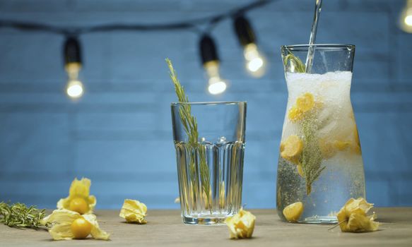 Close up sparkling water pouring into the glass jug with rosemary and physalis. Making of cold drink. Blurry lights on blue background