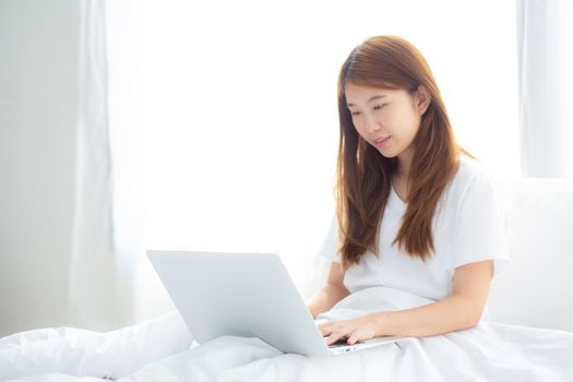 Portrait of beautiful asian young woman setting on bed using laptop computer at bedroom for leisure and relax, freelance with girl working notebook, communication and lifestyle concept.