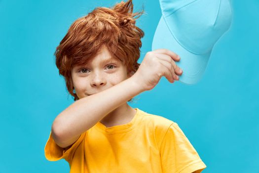 Red-haired boy with freckles holds a cap in his hands in a yellow t-shirt blue isolated fund