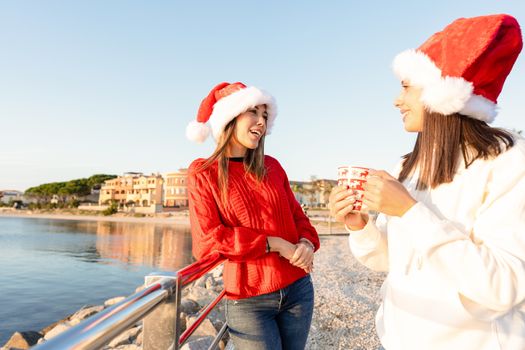 Beautiful young caucasian women standing outdoor talking with a Christmas mug in hands wearing Santa hat - Sunset sea winter vacation for two female friends at Golfo Aranci in Costa Smeralda, Sardinia