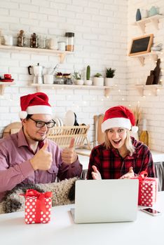 Christmas online greetings. Excited young couple in santa hats greeting their friends in a video call on laptop sitting at the kitchen