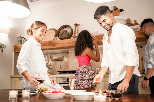 Handsome millennials friends group bonding at home preparing snacks for aperitif - Two heterosexual couple having fun together in the kitchen cooking for dinner - Focus on bearded man with white shirt