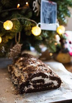 Traditional Christmas chocolate roll with cream on a background of Christmas tree lights on a dark background. View from above
