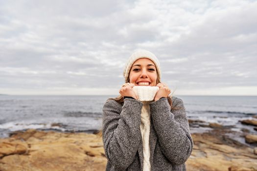 Beautiful young Caucasian woman tightens the neck of her white sweater with her hands for the cold winter weather smiling looking at the camera on sea rocks of Sardinia vacation resort with cloudy sky