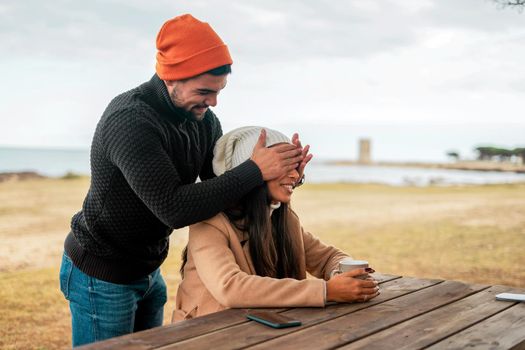 Young heterosexual couple having fun outdoor in sea resort wearing fall-winter clothes and wool hats - Handsome smiling guy blindfolds from behind with hands his girlfriend sitting with a tea cup