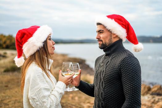 Beautiful heterosexual couple in love toasting outdoor wearing Santa Claus hat looking into each other's eyes holding white wine glass in hand with sea or ocean background in selective focus