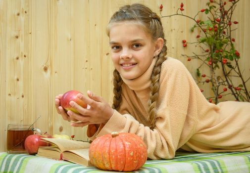 A girl lying down reads a book, holds an apple in her hands and looks happily into the frame