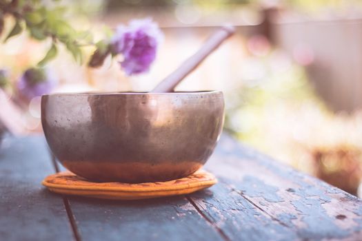 Metal singing bowl on a rustic green, wooden table outdoors. Flowers in the colourful, blurry background