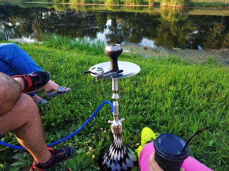 A company of friends smokes a hookah in a green park by the river. Fun with friends