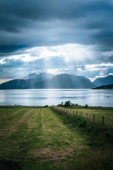 Mystic landscape lake scenery in Scotland: Cloudy sky, meadow, trees and lake with sunbeams, mountain range in the background. Loch Linnhe. 