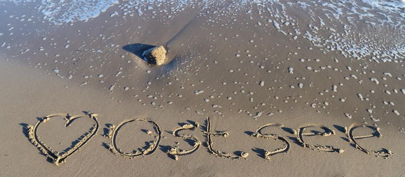 The german word Ostsee (Baltic Sea) and a heart written into the sand of the beach.
