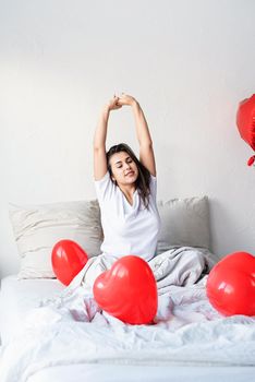 Young happy brunette woman sitting awake in the bed with red heart shaped balloons