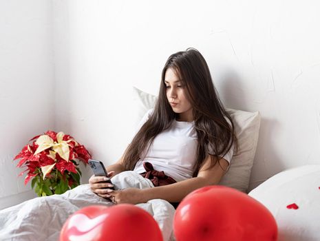 Young brunette woman sitting awake in the bed with red heart shaped balloons and decorations texting