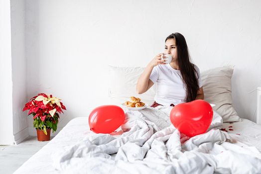 Young brunette woman sitting awake in the bed with red heart shaped balloons and decorations drinking coffee eating croissants
