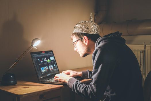 Conspiracy theory concept: young man with aluminum cap searching the internet, sitting lonely in the dark basement 