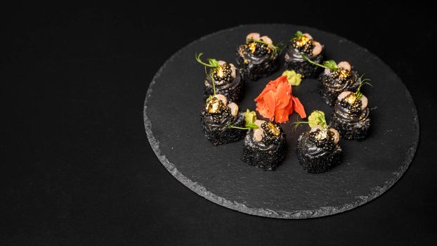 Sushi roll with nori, black rice, crab meat, cucumber, avocado, smoked salmon mousse, oar caviar, masago, shrimp cocktail and edible gold leaf with red ginger and wasabi on black table and background.