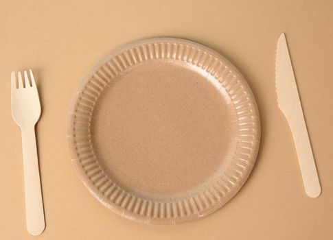 wooden fork and empty round brown disposable plate made from recycled materials on a brown background, top view. Concept of the absence of non-recyclable garbage, rejection of plastic, top view