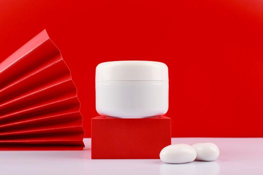 Close up of white glossy jar with cream, mask or balm on podium against red background decorated with stones and waver