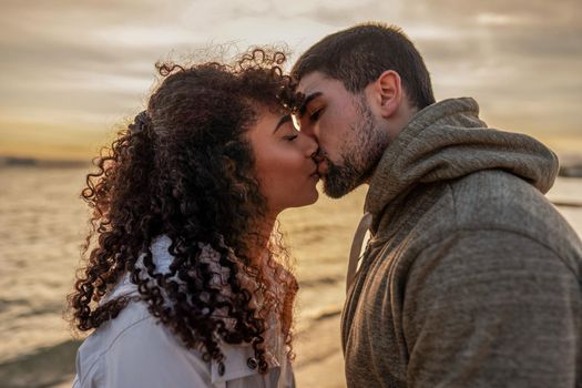 Head and shoulders portrait of young beautiful couple in love kissing at sunset in winter seaside resort with cloudy sky. Two millennials in vacation travel manifesting their heart sentiments outdoor