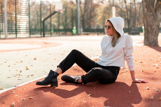 Outdoor portrait of young beautiful woman with long in sunglasses and a white hooded sweater sitting on the sportsground track