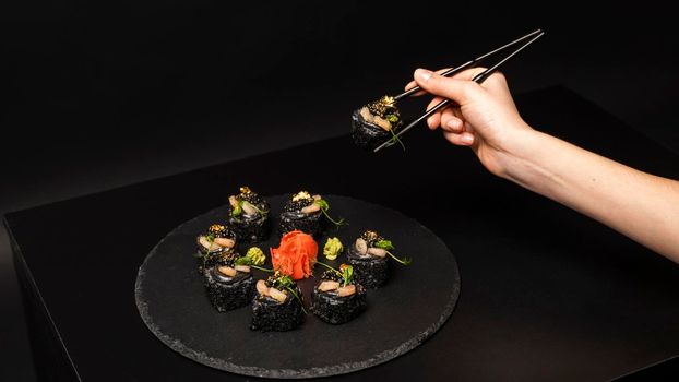 Hand with chopsticks holds a sushi. Custom sushi roll with black rice, crab meat, avocado, smoked salmon mousse, oar caviar, masago, shrimp cocktail, edible gold leaf, ginger, wasabi on black table.