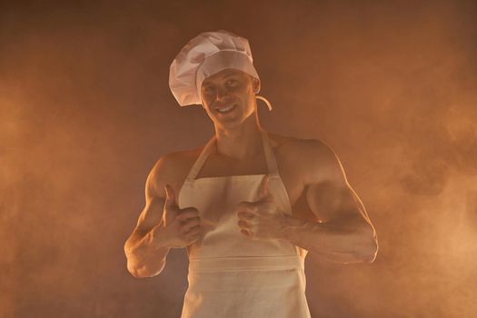Portrait of muscular chef wearing white apron and chef hat, showing thumb up on smoky background