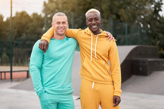 International friendship. Two happy diverse men in bright sportsuits standing outdoor, bonding together and smiling to camera in urban park