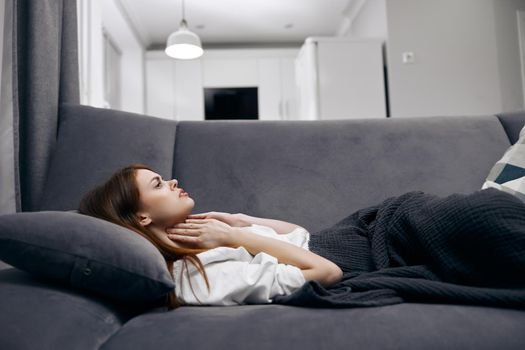 woman holding her neck while lying on the couch hiding with a blanket at home feeling unwell