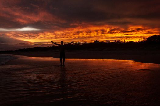 young man rising hands in the air because he is happy, with a colorfull sunset in the background at Noosaville beach, Sunshine Coast, Australia.