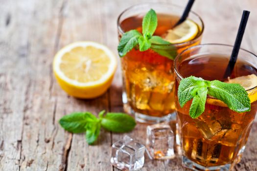 Cold iced tea with lemon, mint leaves and ice cubes in two glasses on rustic wooden table background. 