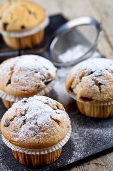 Two fresh homemade muffins with sugar powder and metal strainer closeup on black board.
