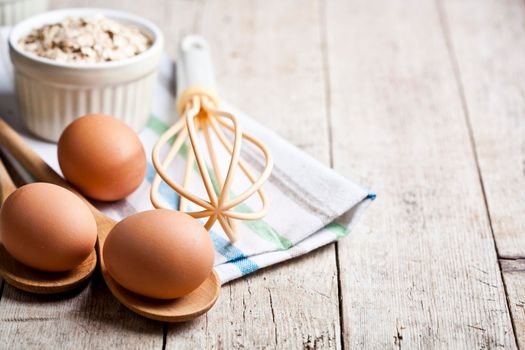 Fresh chicken eggs, oat flakes in ceramic bowl and kitchen utensil on rustic wooden table background.