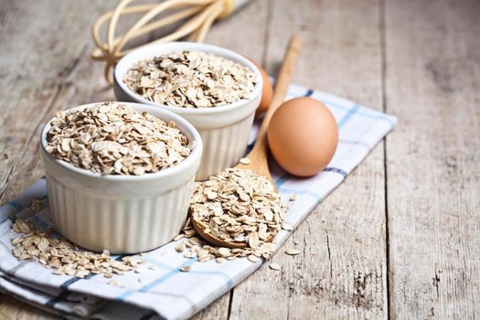 Oat flakes in ceramic bowls and wooden spoon and fresh chicken eggs on rustic wooden table background.