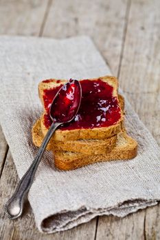 Toasted cereal bread slices stack with homemade cherry jam and spoon closeup on rustic wooden table background. 