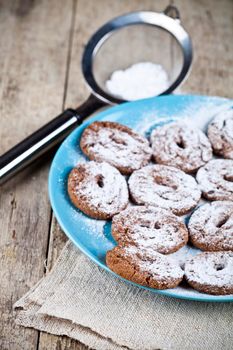 Fresh baked chocolate chip cookies with sugar powder on blue plate and metal strainer closeup on rustic wooden table.