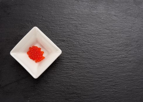 fresh grained red chum salmon caviar in a white ceramic bowl on a black slate stone background