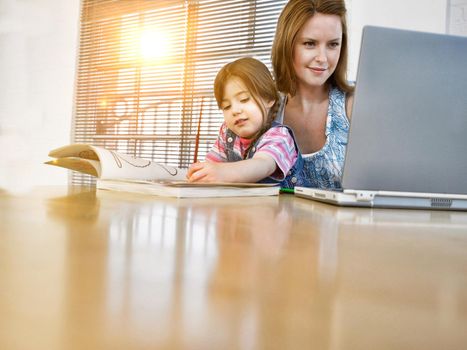 Young woman working with laptop with her daughter colouring on book