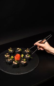 Hand with chopsticks wants to take custom sushi roll with black rice, crab meat, avocado, smoked salmon mousse, oar caviar, masago, shrimp cocktail, edible gold leaf, ginger, wasabi on black table.