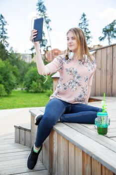 A young girl of 20 years old Caucasian appearance makes a selfie on her mobile phone while sitting on a wooden podium in the park on a summer day.The girl is dressed in a floral T-shirt and jeans, 