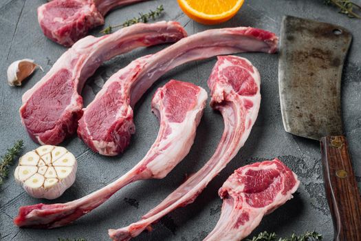 Frenched raw fatty lamb chops, with ingredients carrot orange, herbs, and old butcher cleaver knife, on gray stone background