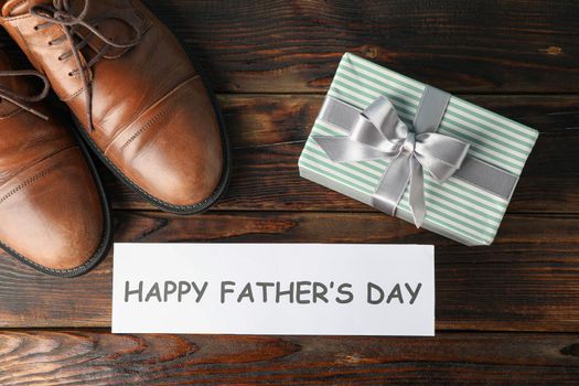 Brown leather shoes, inscription happy fathers day and gift box on wooden background, space for text and top view